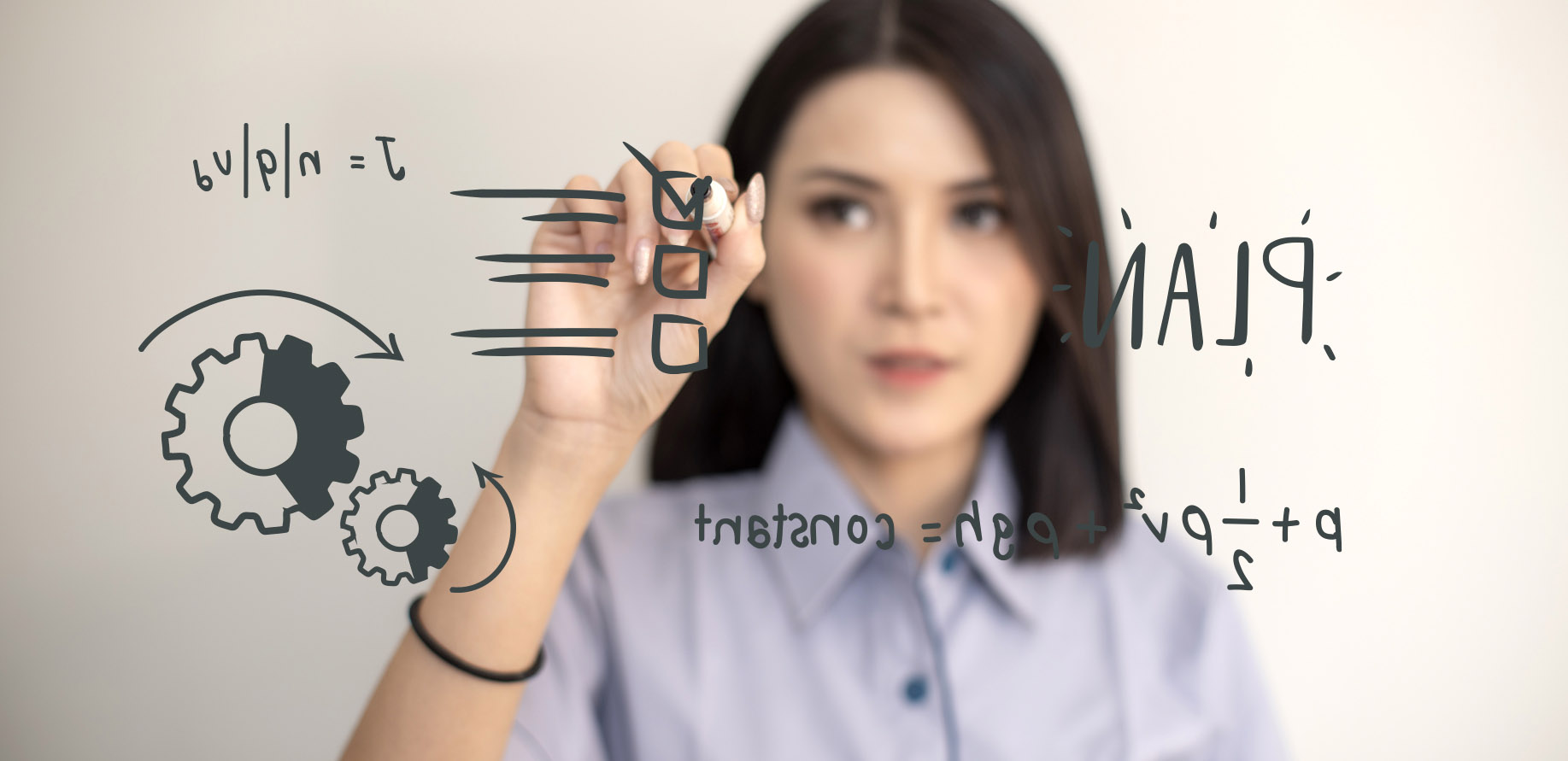 Woman writing on a transparent glass whiteboard filled with sketches of gears and formulas