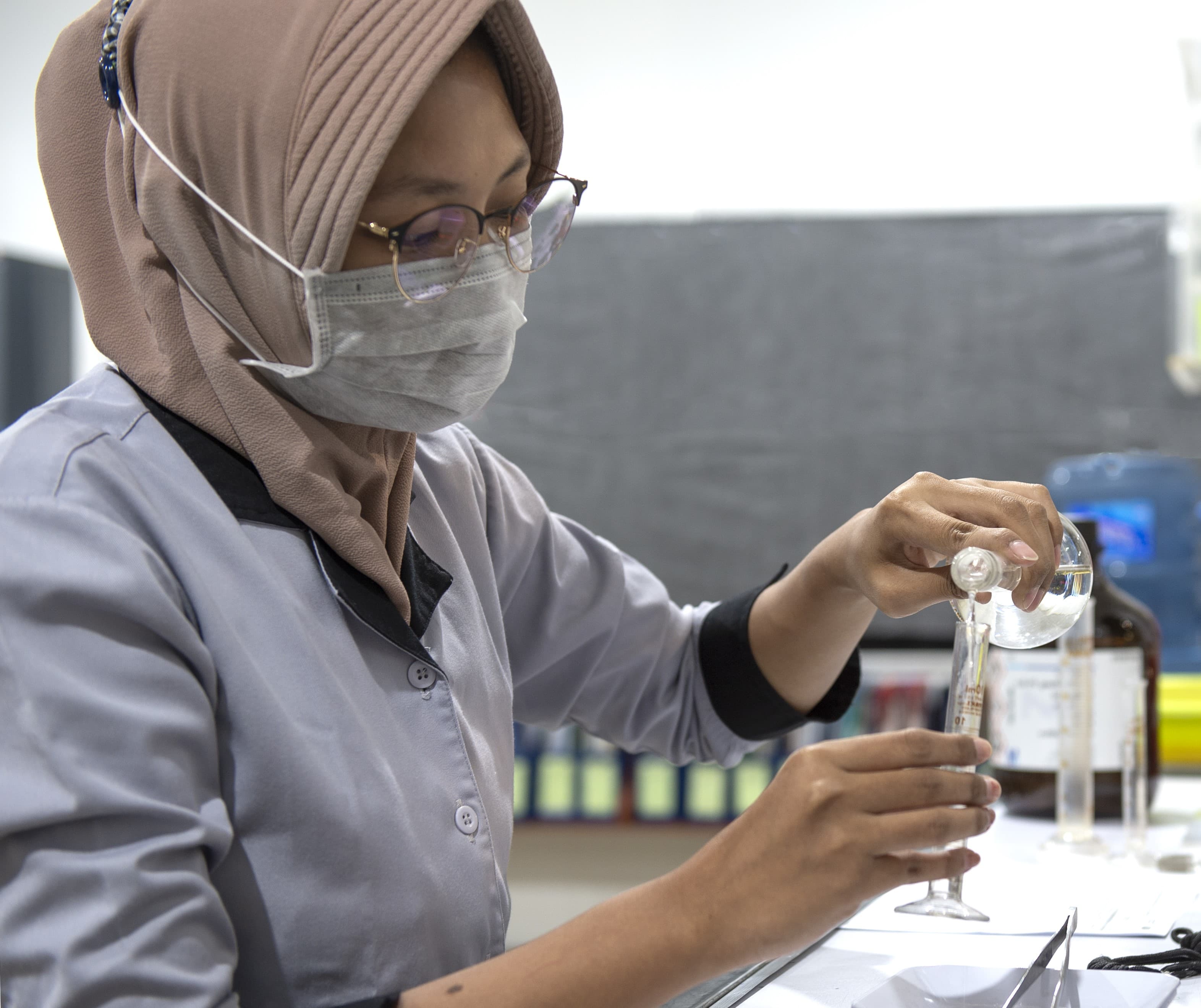 Female laboratory technician wearing a hijab and face mask pouring liquid into a test tube