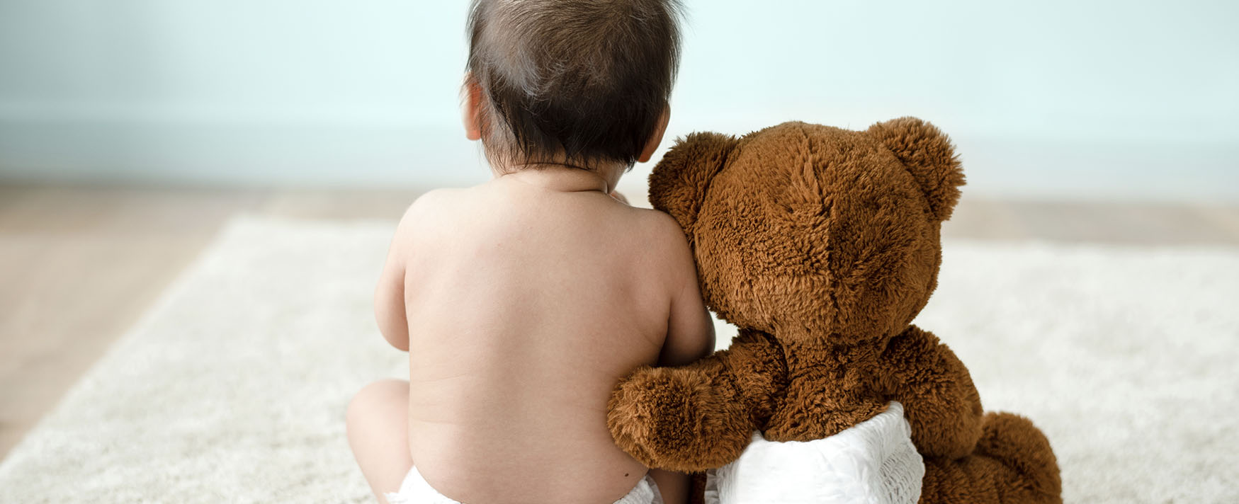 Baby wearing a white diaper sitting on a fluffy rug next to a teddy bear
