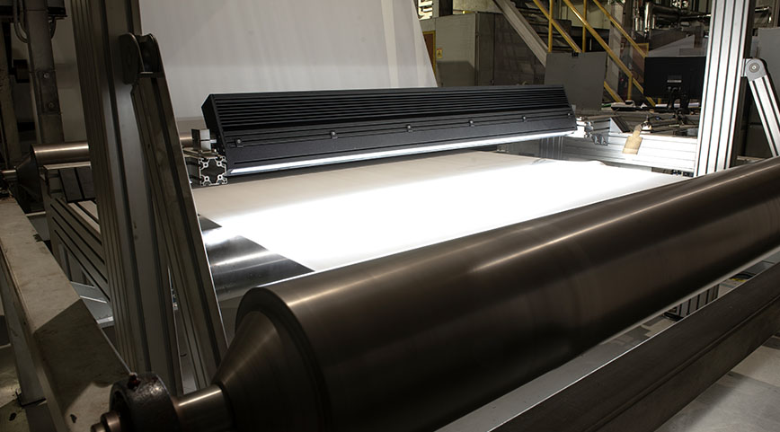 Brightly illuminated white fabric sheet passing under an inspection light on rollers